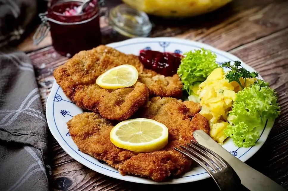 Schnitzel auf der Aldranser Alm in Tirol Das Foto zeigt ein frisch gebackenes Schnitzel mit Zitronenscheiben, serviert mit Kartoffelsalat, grünen Salatblättern und Preiselbeeren auf einem Teller.