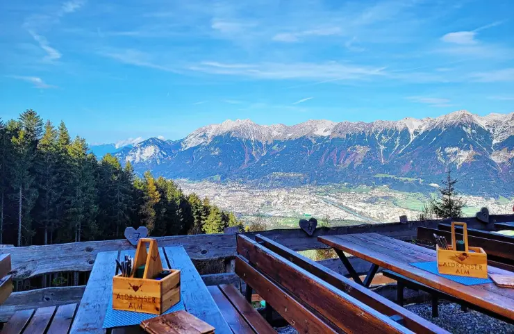 Terrasse der Aldranser Alm mit Panorama Das Foto zeigt die Holzterrasse der Aldranser Alm mit Sitzbänken und Tischen. Dahinter eröffnet sich ein weiter Ausblick über Innsbruck, das Inntal und die markante Bergkette der Nordkette.