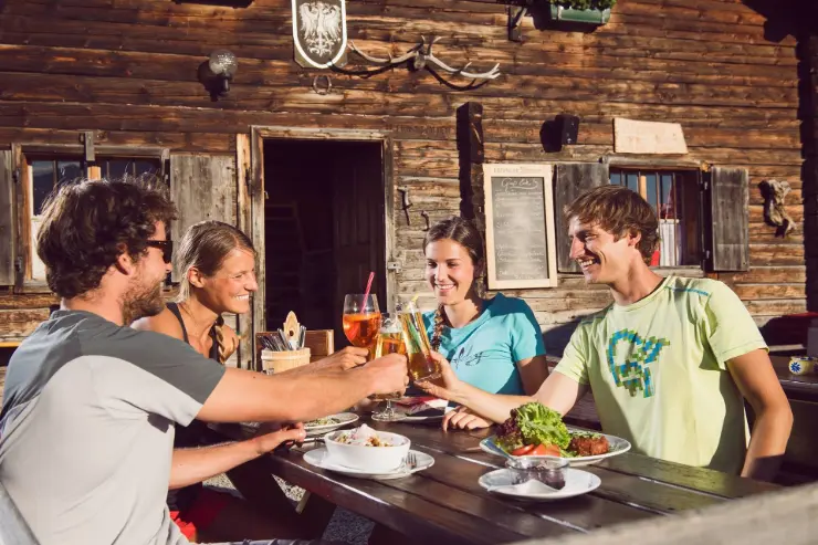 Geselliges Beisammensein auf der Aldranser Alm Das Foto zeigt vier junge Menschen, die auf der Terrasse der Aldranser Alm zusammensitzen, Speisen und Getränke genießen und anstoßen. Im Hintergrund ist die traditionelle Holzhütte der Alm zu sehen.