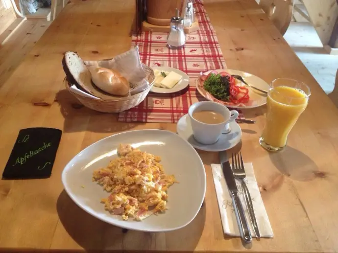 Das Foto zeigt ein typisches Almfrühstück auf der Aldranser Alm: Auf einem Holztisch stehen ein Teller mit Rührei und Speck, ein Brotkorb mit Gebäck, Käse, frisches Gemüse, eine Tasse Kaffee und ein Glas Orangensaft.