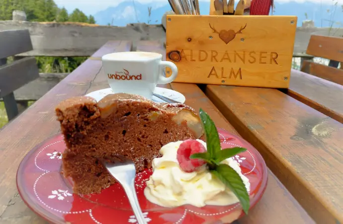 Dessert & Kaffee auf der Aldranser Alm Das Foto zeigt ein Stück Schokoladenkuchen mit Schlagobers und Himbeere, serviert auf einem roten Teller. Daneben steht eine Tasse Kaffee der Marke Unbound. Im Hintergrund sind Holzbänke und eine Box mit der Aufschrift „Aldranser Alm“ zu sehen.